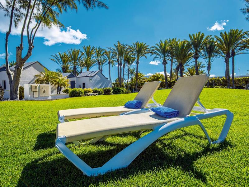 Twee witte ligstoelen met blauwe kussens op groen gras voor palmbomen en huizen.