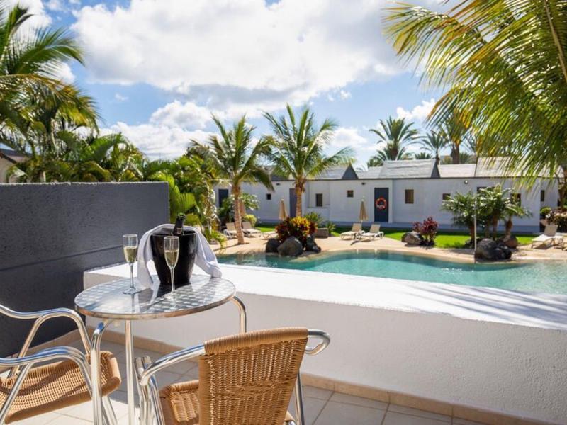 Balkon met tafel en stoelen met uitzicht op zwembad en witte bungalows met palmbomen.