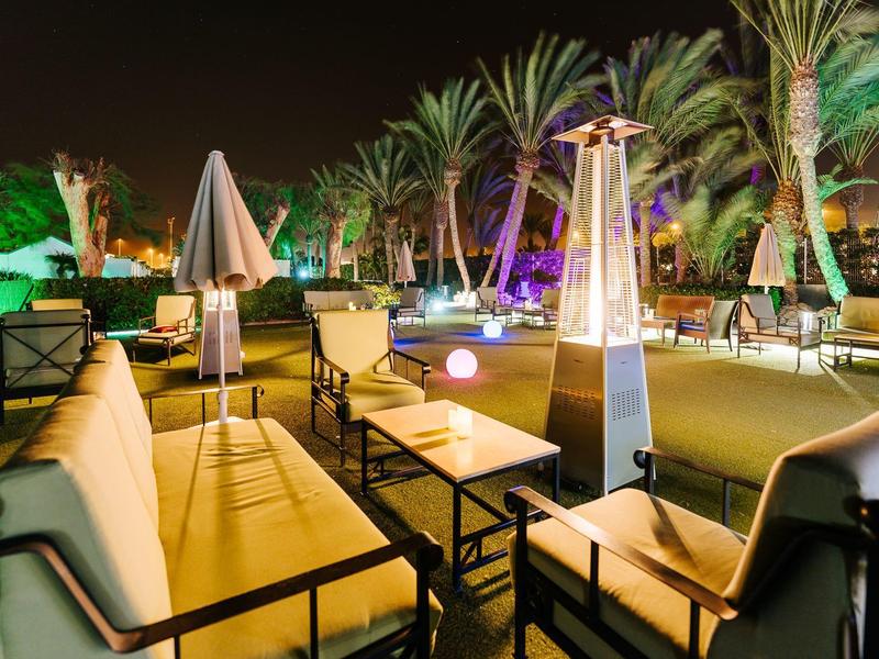 Avond buitenlounge met comfortabele banken, tafels en verwarmde staande lampen omgeven door palmbomen.