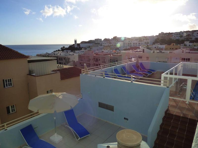 Blick auf eine Terrasse mit zwei blauen Liegestühlen und Sonnenschirm, dahinter Häuser und Meer.