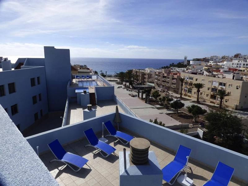 Dachterrasse mit blauen Liegestühlen, runden Tischen und Blick auf Straße und Meer im Hintergrund.