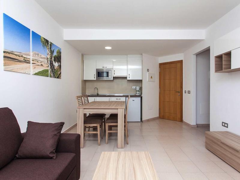 Moderner Wohnraum mit Sofa, Holztisch, Stühlen und offener Küchenzeile in hellem Beige und Weiß.