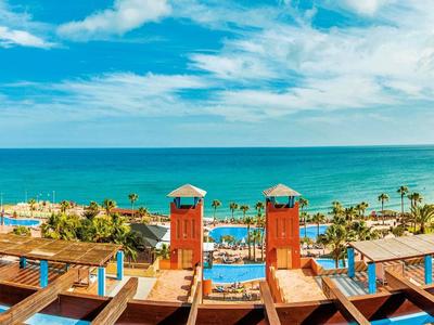 Blick auf ein Hotel mit Pool und Sitzbereich mit Meer im Hintergrund unter blauem Himmel