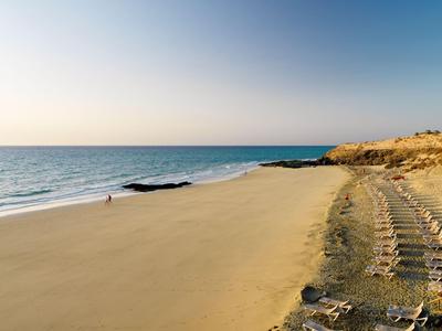 Weiter Sandstrand mit Reihen von Sonnenliegen und einigen Felsen am Ufer bei klarem Himmel.