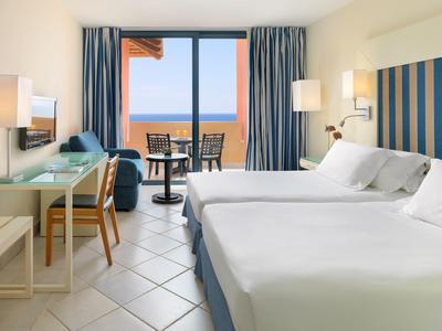 Helles Hotelzimmer mit zwei Einzelbetten, Glas-Schreibtisch, Balkon und Meerblick.