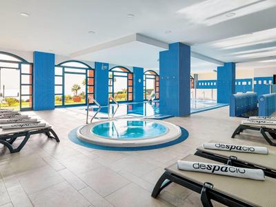 Helles Hallenbad mit blauen Säulen, rundem Whirlpool und schwarzen Liegen mit weißen Handtüchern.