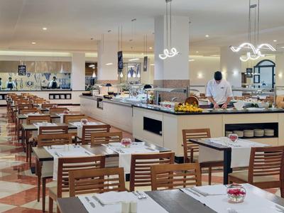 Ein heller und moderner Speisesaal mit Holzstühlen, weißen Tischen und Buffetbereich in einem Hotel.