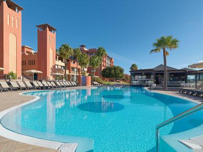 Großer Pool mit Sonnenliegen und Palmen vor roten Hoteltürmen unter blauem Himmel.