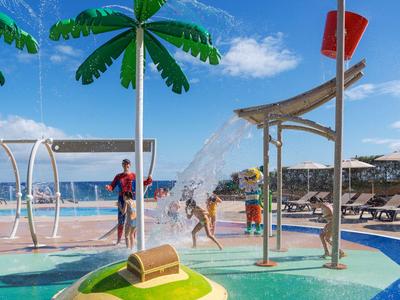 Kinder spielen in einem bunten Wasserspielbereich mit Palmen und Duschen nahe einem Hotelpool.