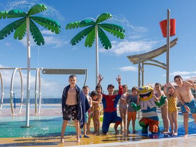Menschen und eine kostümierte Figur posieren am Pool mit Palmen vor dem Meer an einem sonnigen Tag.