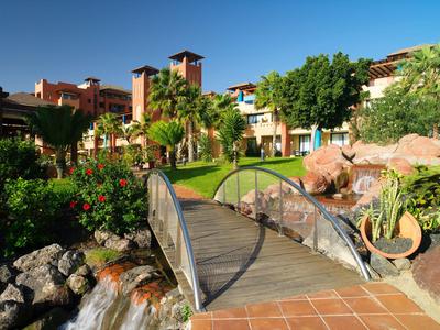 Hotelgarten mit Holzbrücke, üppiger Vegetation und blauem Himmel im Hintergrund.