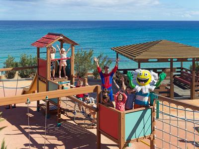 Kinder spielen fröhlich auf einem bunten Spielplatz am Meer mit einem Pavillon im Hintergrund.