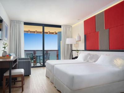 Helles Hotelzimmer mit Doppelbett, Schreibtisch, Sesseln und Balkon mit Meerblick.