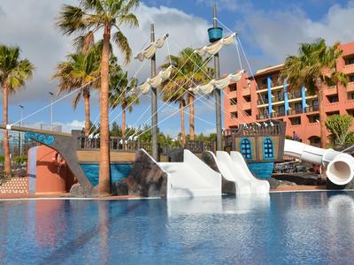Wasserrutschen und Pool mit Palmen vor einem rosa Hotel unter blauem Himmel.