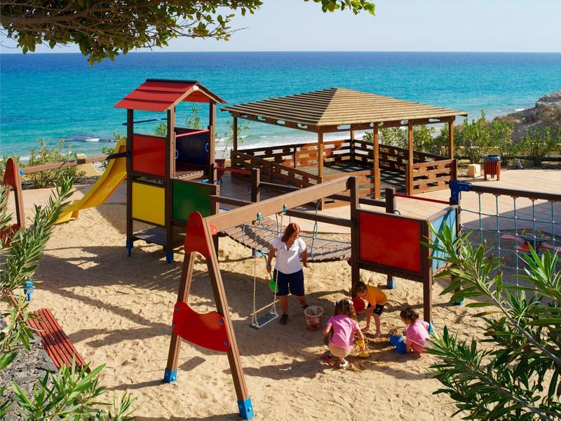Bunter Spielplatz mit Klettergerüst und Sand, Kinder spielen, dahinter Meer und blauer Himmel.