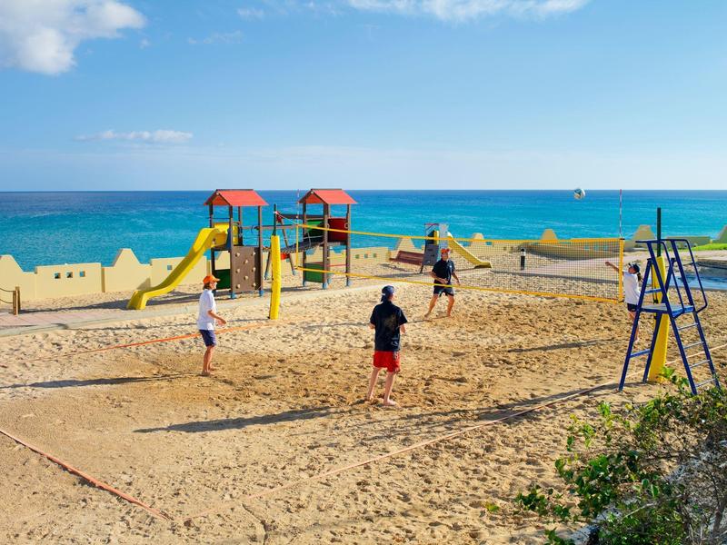 Spielende Personen auf Sandstrand-Volleyballfeld mit blauer See und klarem Himmel im Hintergrund.