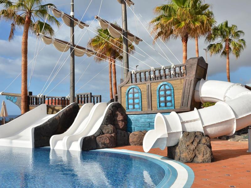 Wasserpark mit mehreren weißen Rutschen, Palmen und einem großen Schiff als Deko bei sonnigem Himmel.