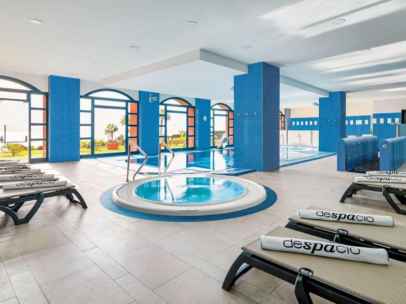 Helles Hallenbad mit blauen Säulen, rundem Whirlpool und schwarzen Liegen mit weißen Handtüchern.