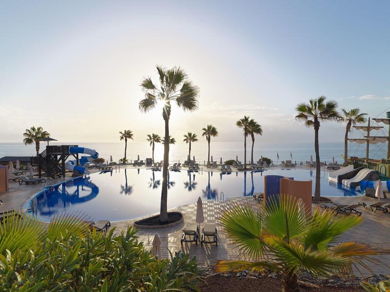 Großer Pool mit Palmen und Sonnenschirmen, blauem Himmel und Sonnenlicht im Hintergrund.