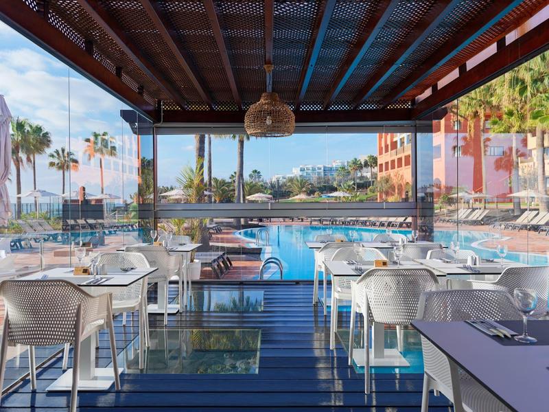 Offenes Restaurant mit weißen Stühlen, Holzdecke und Blick auf blauen Pool und Palmen.
