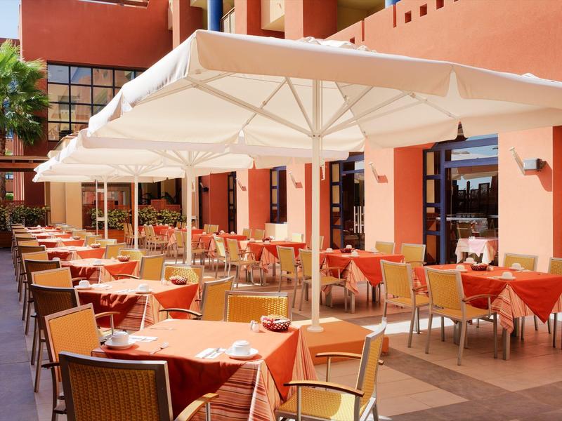 Freiluftrestaurant mit orangenen Tischtüchern, gelben Stühlen und großen weißen Sonnenschirmen.