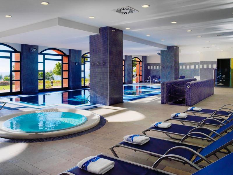 Moderner Innenpool mit Whirlpool, blauen Liegestühlen und großen Fenstern mit Meerblick.