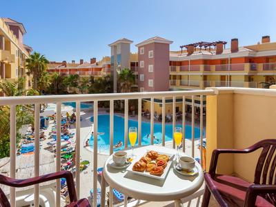 Balkon mit kleinem weißen Tisch, zwei Stühlen und Blick auf einen Pool und bunte Hotelgebäude.