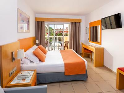 Helles Hotelzimmer mit Doppelbett, orangefarbener Bettdecke, TV und Balkon mit Blick ins Grüne.