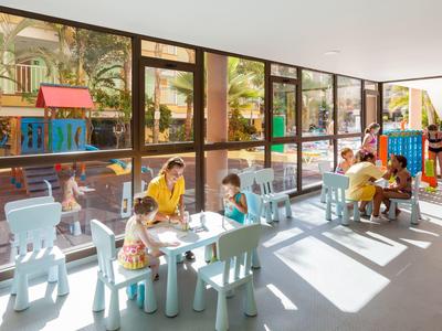 Heller Spielraum mit großen Fenstern, Kindertischen und spielenden Kindern sowie Betreuern.