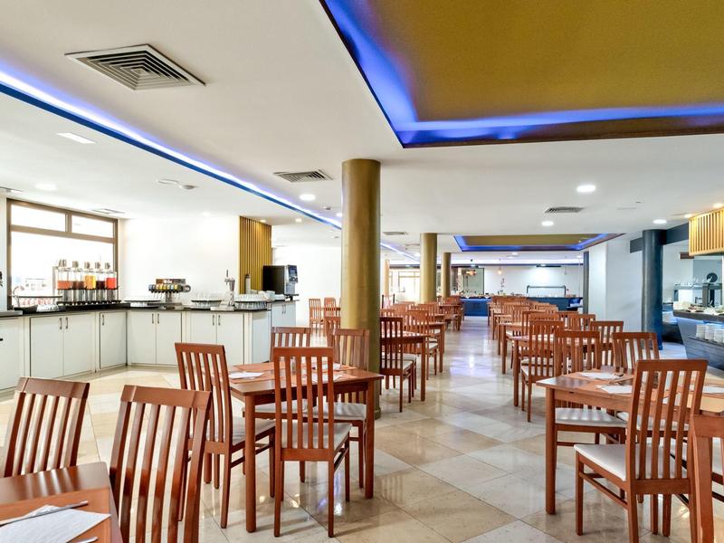 Großer, heller Speisesaal mit vielen hölzernen Stühlen und Tischen, hellen Wänden und blauen Deckenlichtern.