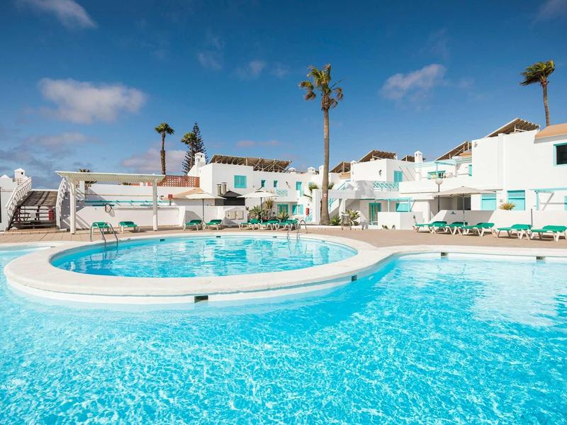 Blue pool with white loungers in front of white hotel buildings under a blue sky.