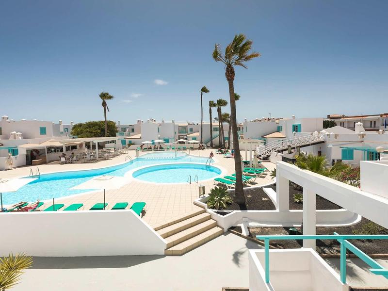 A modern outdoor pool with white buildings and palm trees under a clear sky.