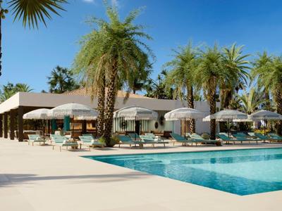 Outdoor pool with sun loungers and umbrellas surrounded by palm trees at a sunny resort.