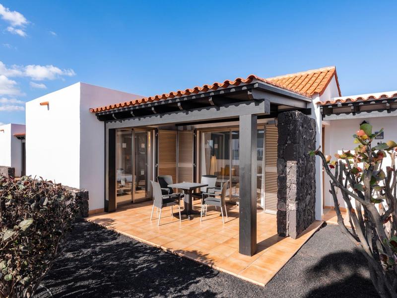 Modern bungalow with terrace, large glass windows, and seating area under sunny weather.