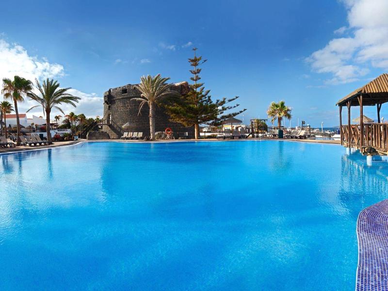 Large outdoor pool with sun loungers and palm trees under blue sky in tropical resort.