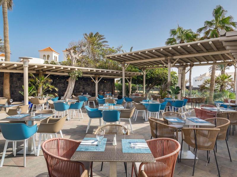 Open-air restaurant with wooden and wicker furniture, tables set with blue placemats, surrounded by palm trees.