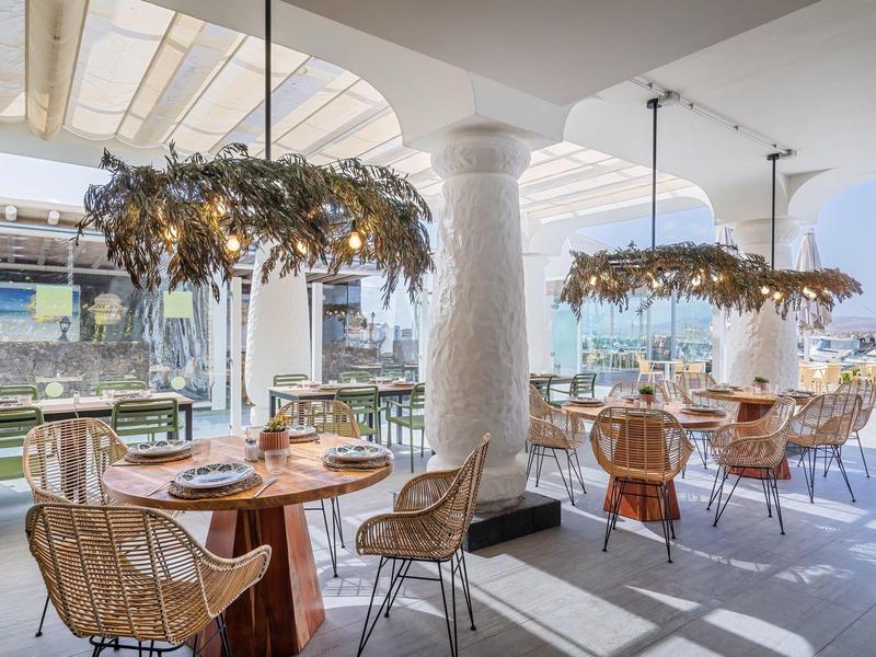 Bright restaurant with wooden tables, wicker chairs, decorated chandeliers, plenty of daylight.