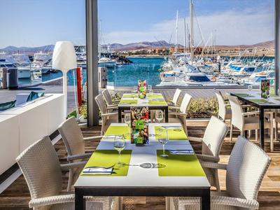 Restaurantterrasse mit weißen Stühlen und Tischen, Blick auf Hafen und Boote bei sonnigem Wetter.