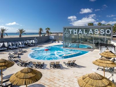 Ein Wellnessbereich mit Pool, Liegestühlen und Palmen vor dem Meer bei sonnigem Wetter.