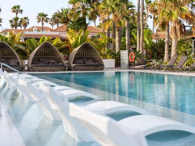 Moderner Poolbereich mit weißen Liegestühlen, Palm Bäumen und schattigen Tagesbetten im Hintergrund.