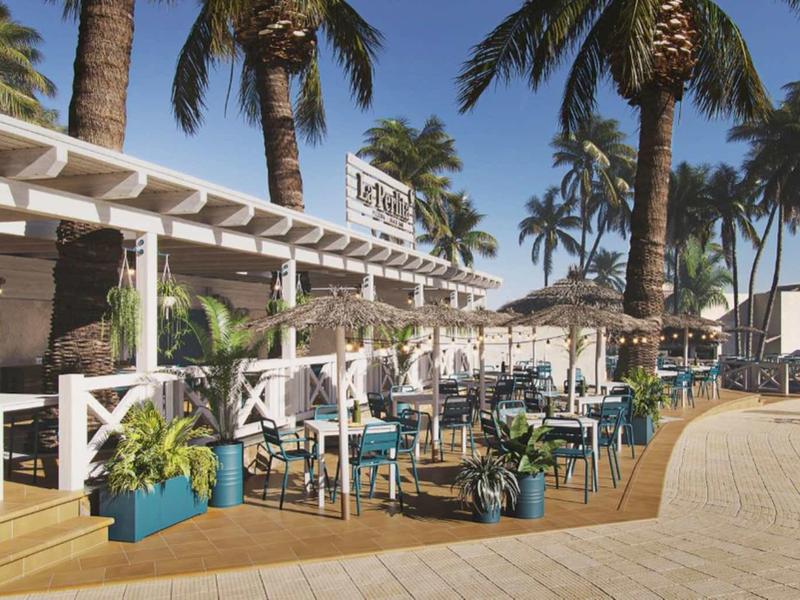 Offenes Strandcafé mit weißen Möbeln, blauen Stühlen und Palmen an einem sonnigen Tag.