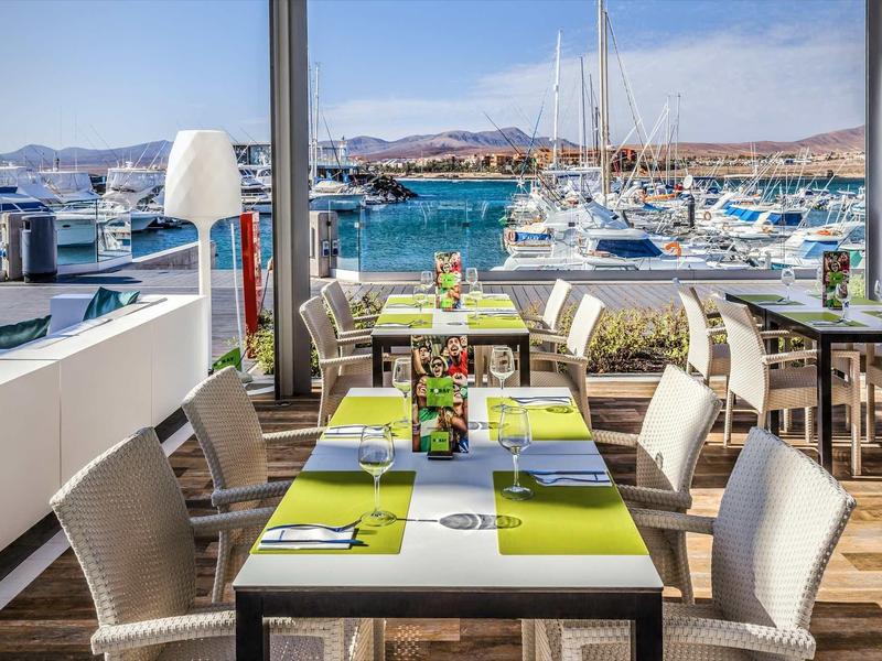 Restaurantterrasse mit weißen Stühlen und Tischen, Blick auf Hafen und Boote bei sonnigem Wetter.