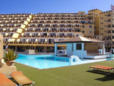 Großes Hotel mit vielen Balkonen und einem hellblauen Pool in der Mitte, umgeben von Liegestühlen.