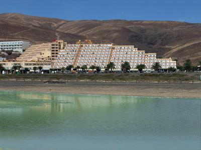 Großes terrassenförmiges Hotelgebäude vor bergiger Landschaft mit grünem Wasser im Vordergrund.