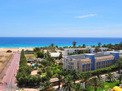 View of a hotel with pool and palm trees by a sunny beach under a blue sky.