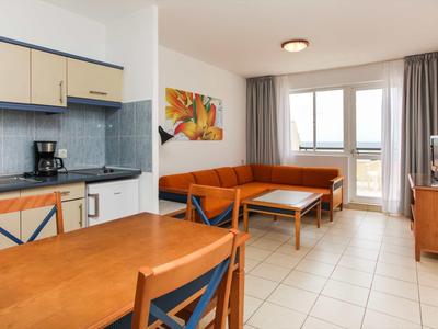 Bright living room with orange sofa, dining table, and kitchenette in a hotel room.