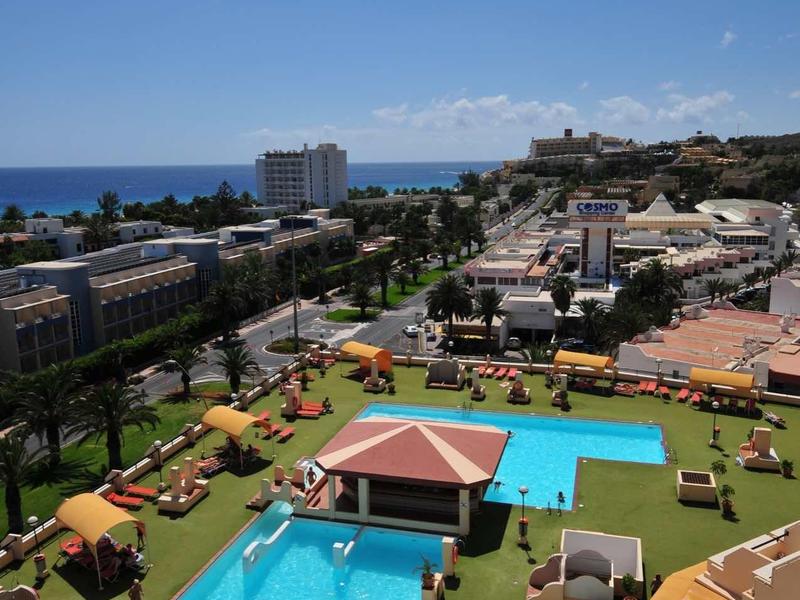 View of hotel pool with sun loungers, buildings, and sea in the background under clear sky