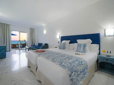 Bright hotel room with two beds, blue headboard, and access to a balcony with sea view.