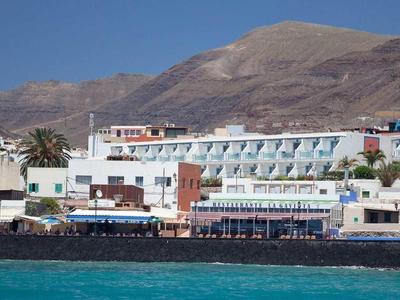 Coastal town with hotels, white buildings, and a mountainous backdrop by clear blue water.