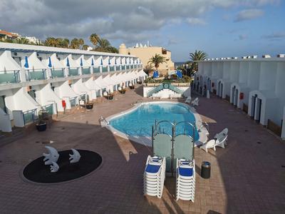 A hotel courtyard with a central swimming pool, lounge chairs, and adjacent white buildings under a cloudy sky.
